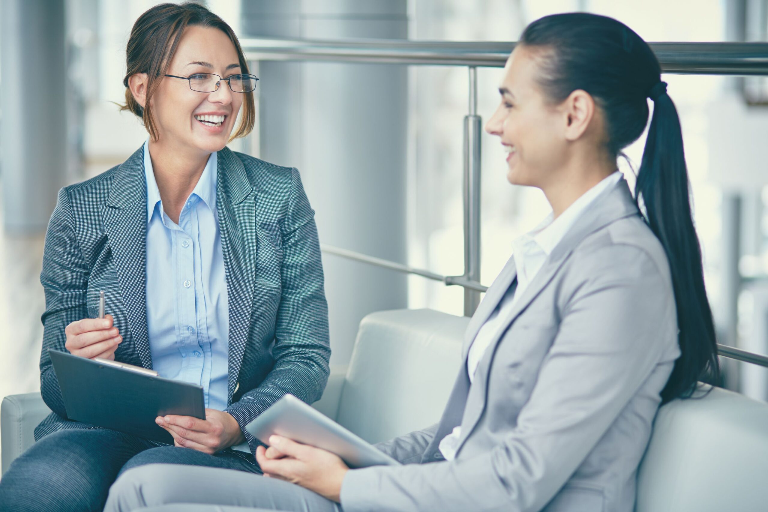 Two female colleagues having a positive workplace conversation while holding a clipboard and tablet